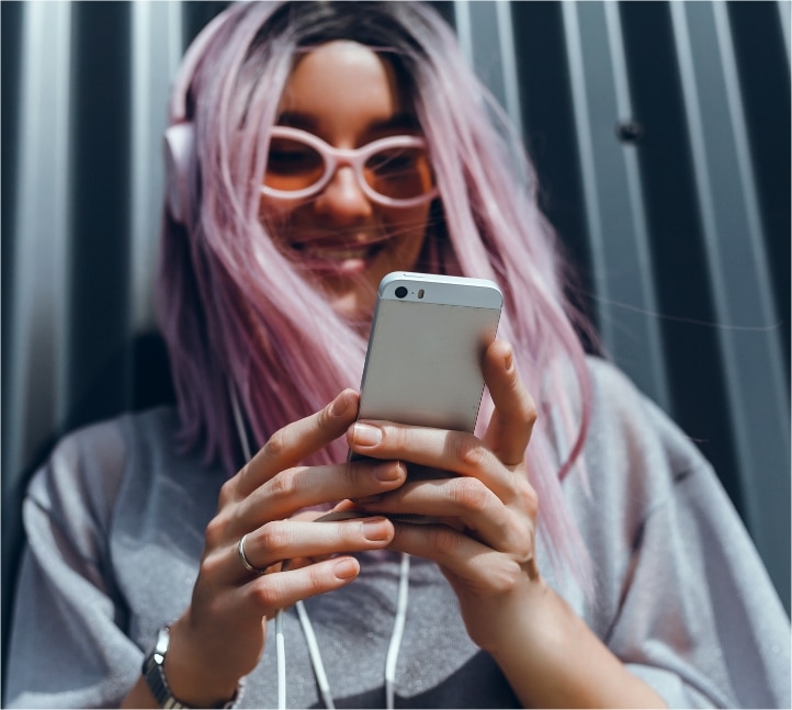 Na zdjęciu widać młodą, uśmiechniętą kobietę z różowymi włosami i okularami, ubrana w szarą bluzę. Kobieta ma założone słuchawki i trzyma w rękach smartfon. Tło stanowi metalowa ściana z pionowymi panelami.