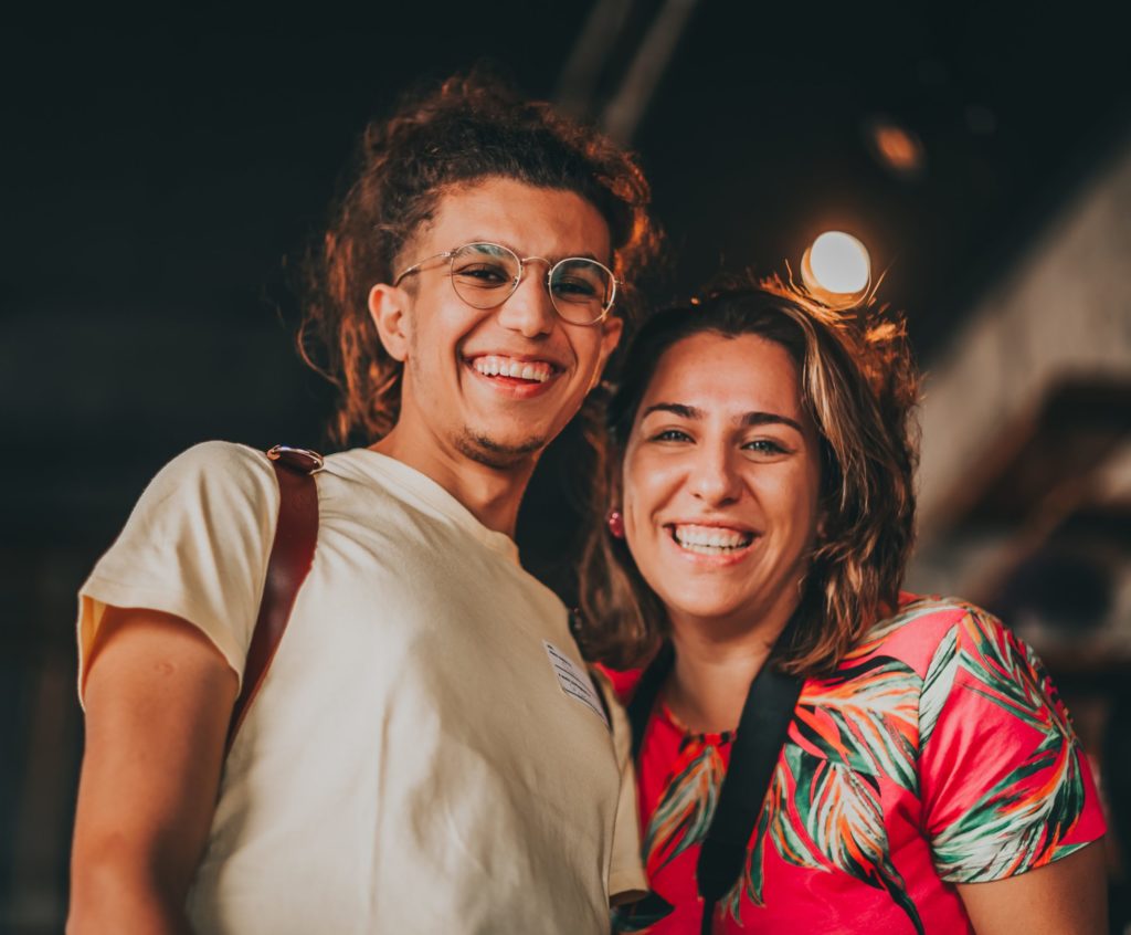 Zdjęcie przedstawia uśmiechniętą parę młodych ludzi stojących blisko siebie. Osoba po lewej stronie ma okulary, długie, kręcone włosy związane w kucyk oraz jasną koszulkę i plecak na ramieniu. Osoba po prawej stronie ma krótsze włosy, kolorową, wzorzystą koszulkę i aparat fotograficzny na pasku.