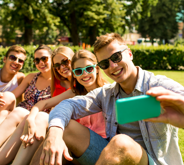 Na zdjęciu widzimy grupę pięciu osób siedzących na trawie w słoneczny dzień, prawdopodobnie w parku. W tle widać drzewa i krzewy. Jedna z osób trzyma zielony smartfon i wygląda na to, że robi selfie. Wszyscy ubrani są w letnie, swobodne stroje, co podkreśla luźną, wakacyjną atmosferę. Wyraz twarzy i gesty wskazują na przyjacielską relację i miłe spędzanie czasu.
