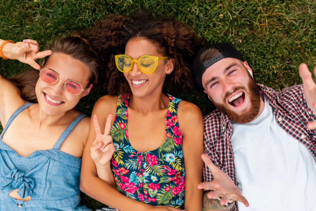 Na zdjęciu widzimy trójkę młodych, uśmiechniętych osób leżących na trawie. Po lewej znajduje się kobieta w jasnych okularach przeciwsłonecznych i niebieskiej sukience, pokazująca znak „peace”. W środku kobieta z kręconymi włosami, w żółtych okularach i kolorowej sukience w kwiaty, również uśmiechnięta. Po prawej stronie leży mężczyzna z brodą, czapką z daszkiem do tyłu, w koszuli w kratę i białej koszulce – szeroko się uśmiecha.