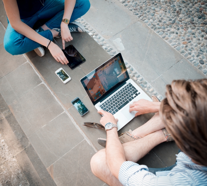 Obraz przedstawia dwie osoby siedzące na kamiennej nawierzchni i pracujące z różnymi urządzeniami elektronicznymi. Jedna osoba korzysta z laptopa, a druga interaktywnie używa tabletu. Między nimi, na ziemi, leżą również dwa smartfony.