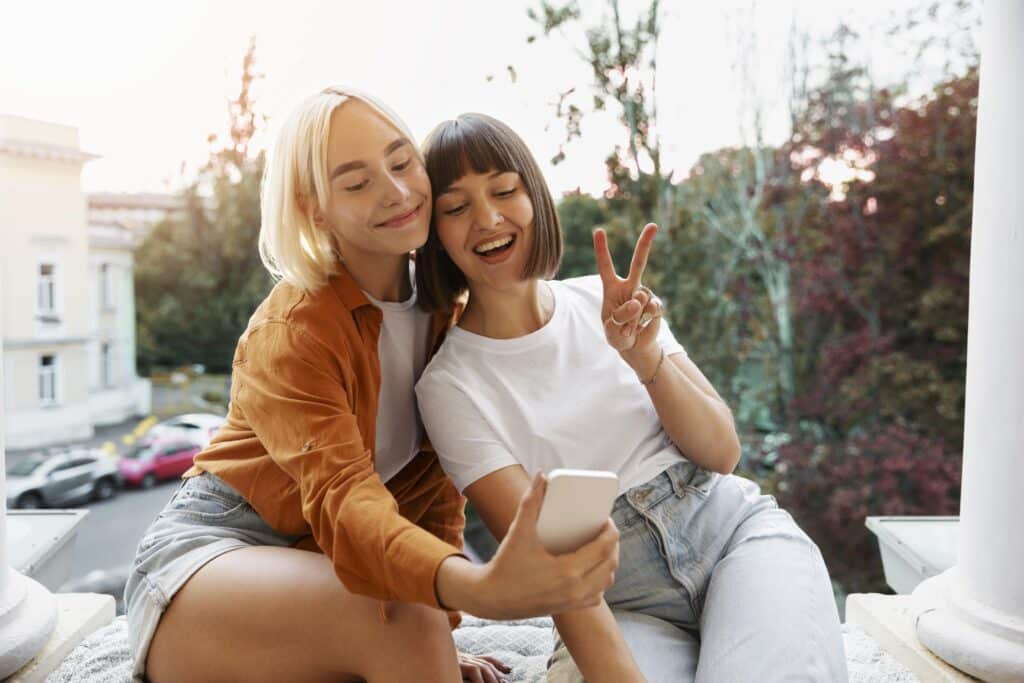 Na zdjęciu widzimy dwie uśmiechnięte kobiety siedzące na zewnętrznej krawędzi balkonu lub tarasu. Jedna z nich ma na sobie białą koszulkę i jasne dżinsy, druga pomarańczową bluzę z długim rękawem oraz szorty. Osoba w białej koszulce pokazuje znak pokoju, a kobieta w pomarańczowej bluzie robi selfie. W tle widać drzewa, budynek i zaparkowane samochody.