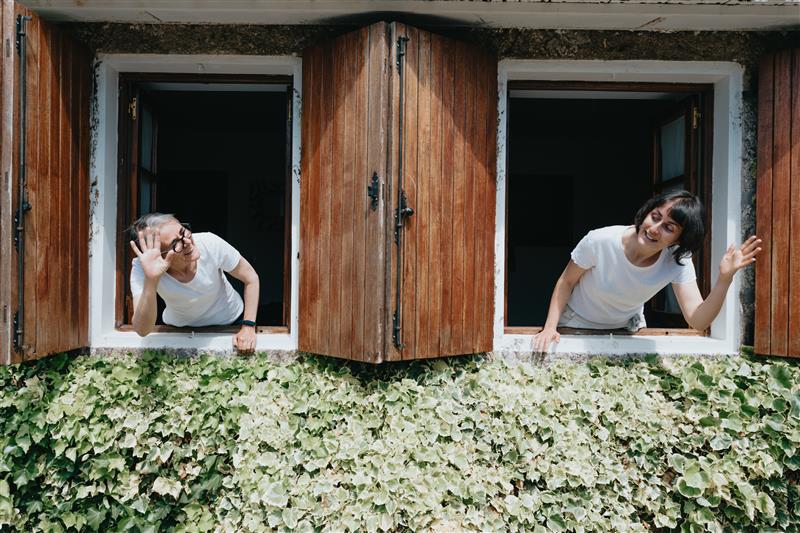 Na zdjęciu widzimy uroczy, porośnięty bluszczem budynek, z którego dwie kobiety wychylają się przez sąsiadujące okna, pozdrawiając się gestem ręki. Obie ubrane są w białe koszulki. Drewniane okiennice podkreślają przytulną, rustykalną atmosferę miejsca.