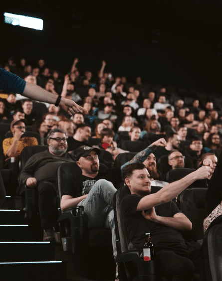 Zdjęcie przedstawia salę kinową pełną widzów. Publiczność siedzi w nowoczesnych, wygodnych fotelach w lekko przyciemnionym oświetleniu. Wiele osób pokazuje coś rękami, śmieje się lub reaguje na to, co dzieje się poza kadrem.